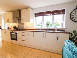 A modern kitchen with white cabinets, a built-in oven, a stainless steel range hood, a window with blinds, and a wall-mounted clock.