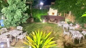 An outdoor patio with white metal furniture arranged around a central stone fire pit. - Corbett Cabana Farm (Haldwani)
