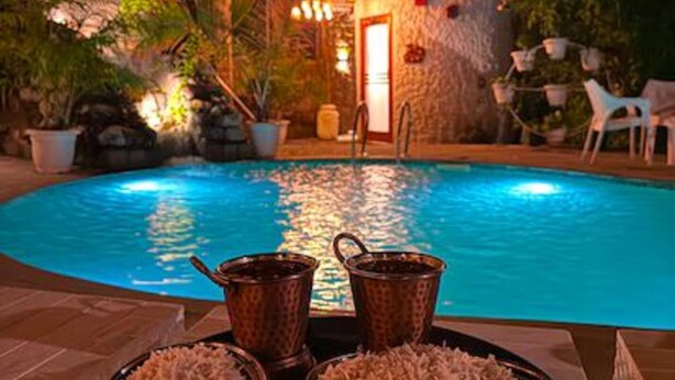 A poolside dining setup with a stone building in the background, surrounded by potted plants and outdoor furniture. - Corbett Cabana Farm (Haldwani)