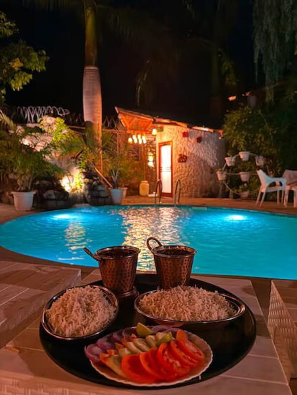 A poolside dining setup with a stone building in the background, surrounded by potted plants and outdoor furniture. - Corbett Cabana Farm (Haldwani)
