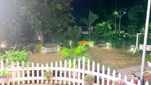 Vista nocturna de un jardín con cerca blanca de piquetes, varias plantas y un letrero amarillo.