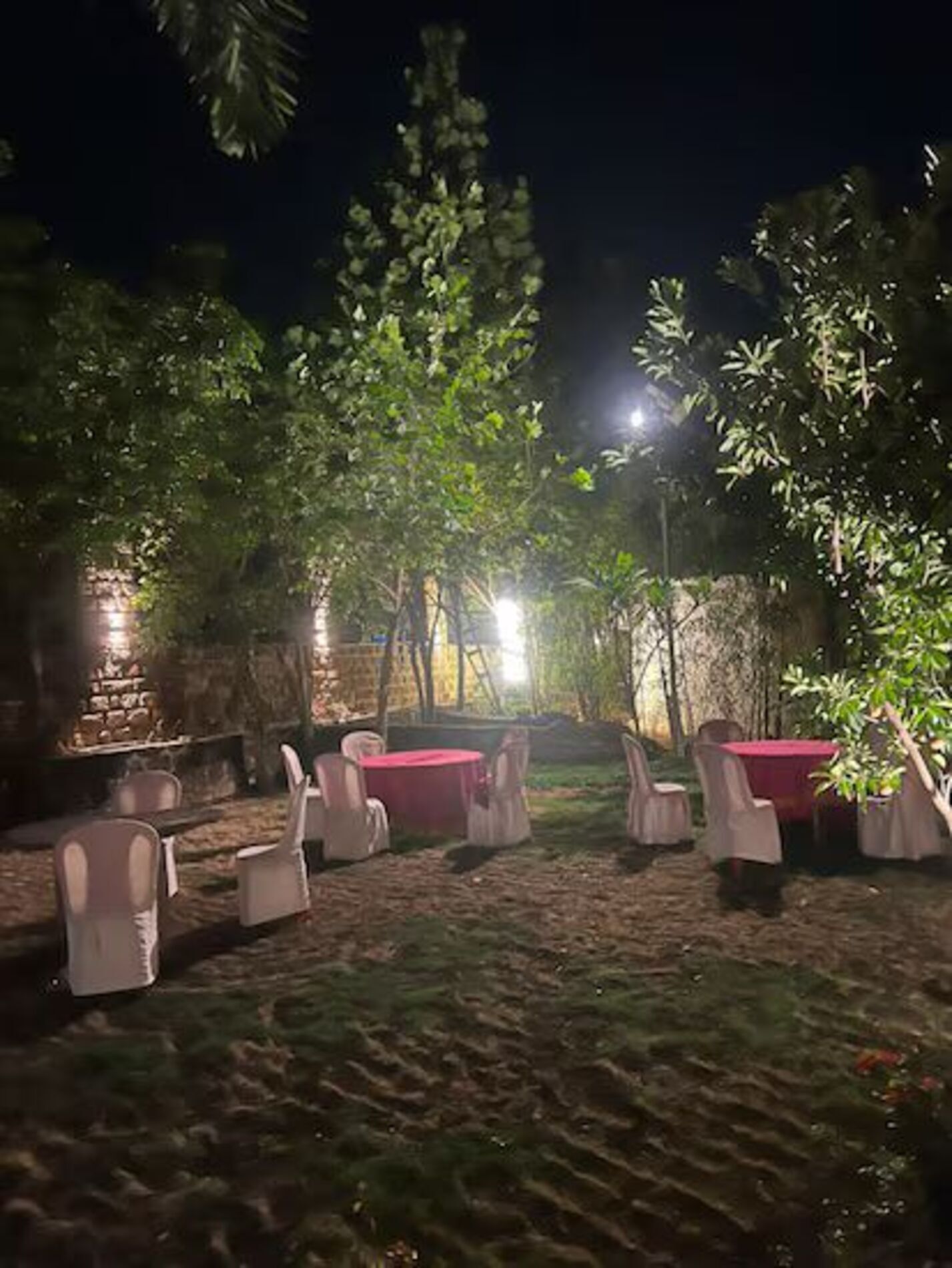 Un espacio al aire libre con mesas y sillas, rodeado de árboles y un muro de piedra.