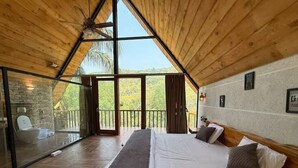 A bedroom with a large bed, wooden ceiling, and a view of trees through large windows. - Corbett Cabana Farm (Haldwani)