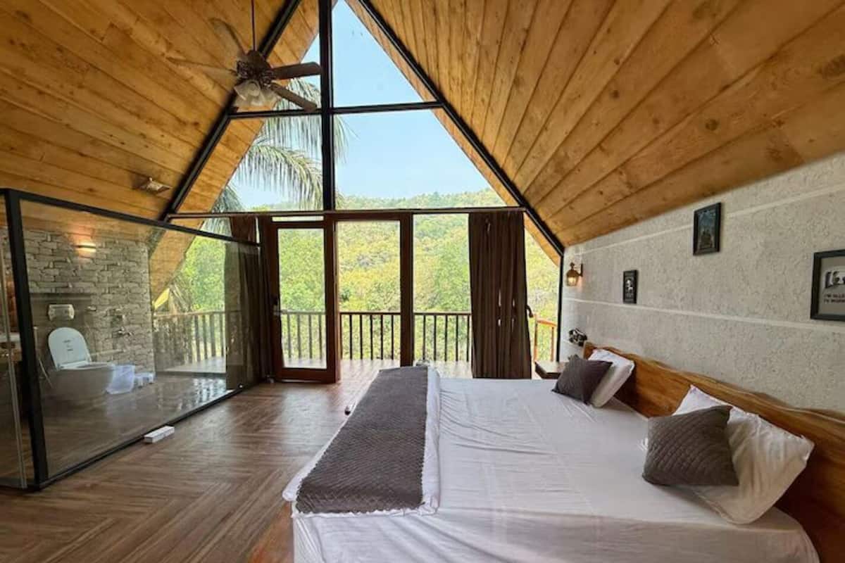 A bedroom with a large bed, wooden ceiling, and a view of trees through large windows.