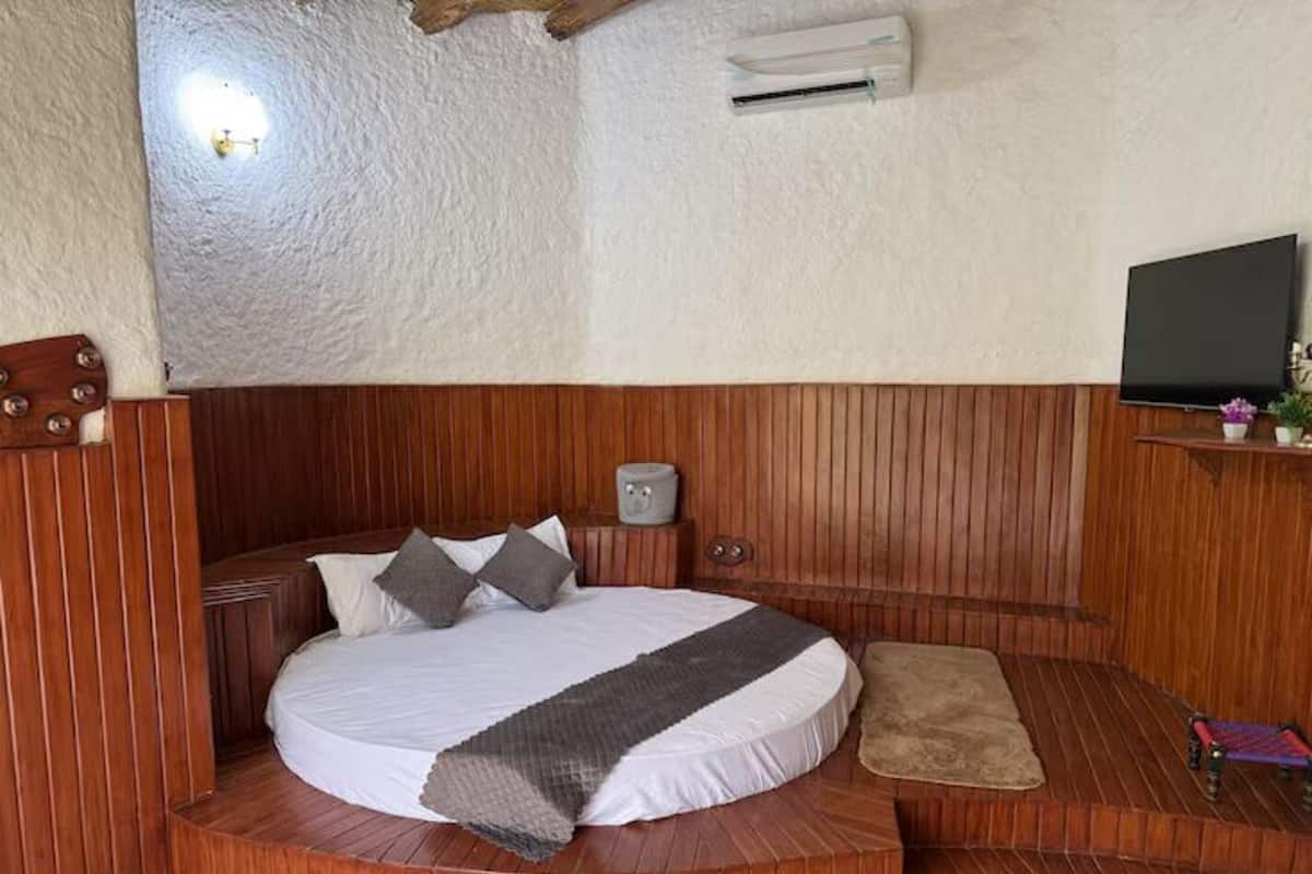 A bedroom with a wooden headboard, a bed with white bedding, a grey blanket, a small bedside table, a wall-mounted air conditioner, and a television on a wooden shelf.