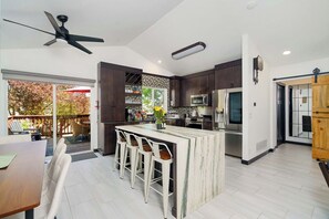 Una cocina moderna con armarios oscuros, una isla central con taburetes de bar, un ventilador de techo y vistas a una terraza exterior.