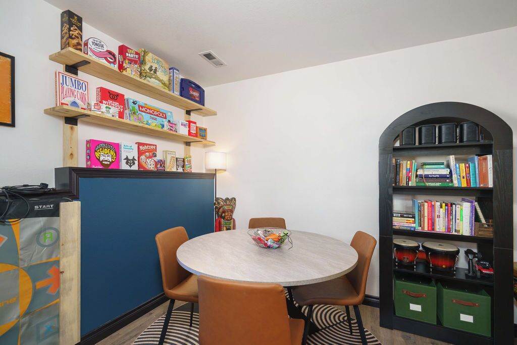 A room with a round table, chairs, a bookshelf, and a desk with a computer.