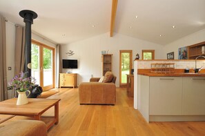 A spacious living area with a sofa, wooden coffee table, and a TV. A kitchen with a wooden countertop and cabinets is visible. - 11 Water's Edge (Looe)