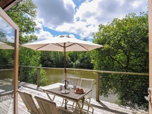 A wooden deck with a table and chairs, an umbrella, and a view of a river surrounded by trees. - 11 Water's Edge (Looe)