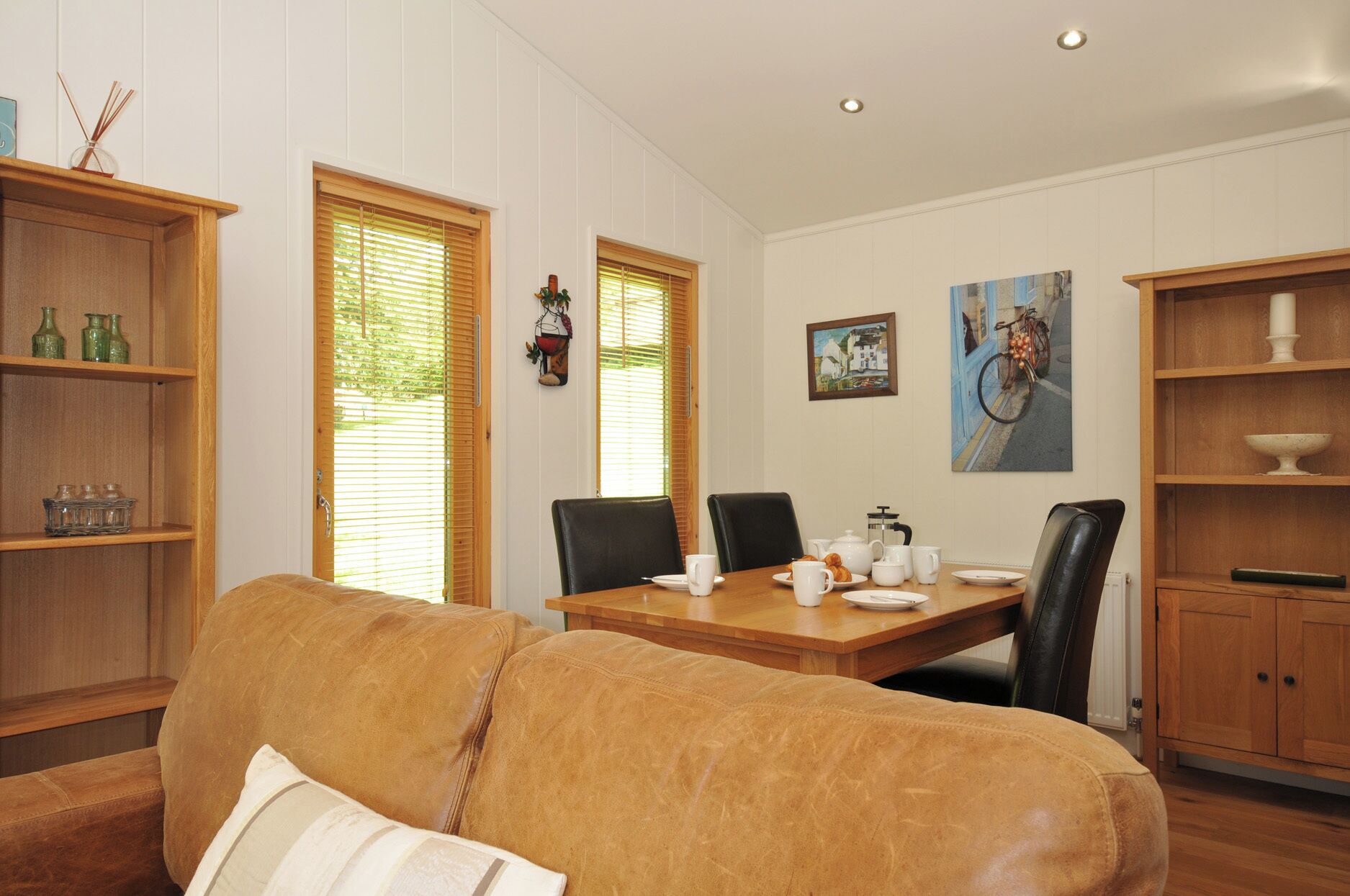Un coin repas avec une table en bois, des chaises et un canapé. On y trouve des étagères en bois ornées d’objets décoratifs et un tableau accroché au mur.