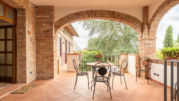 A covered patio with a brick wall, a round table with chairs, and a view of greenery.