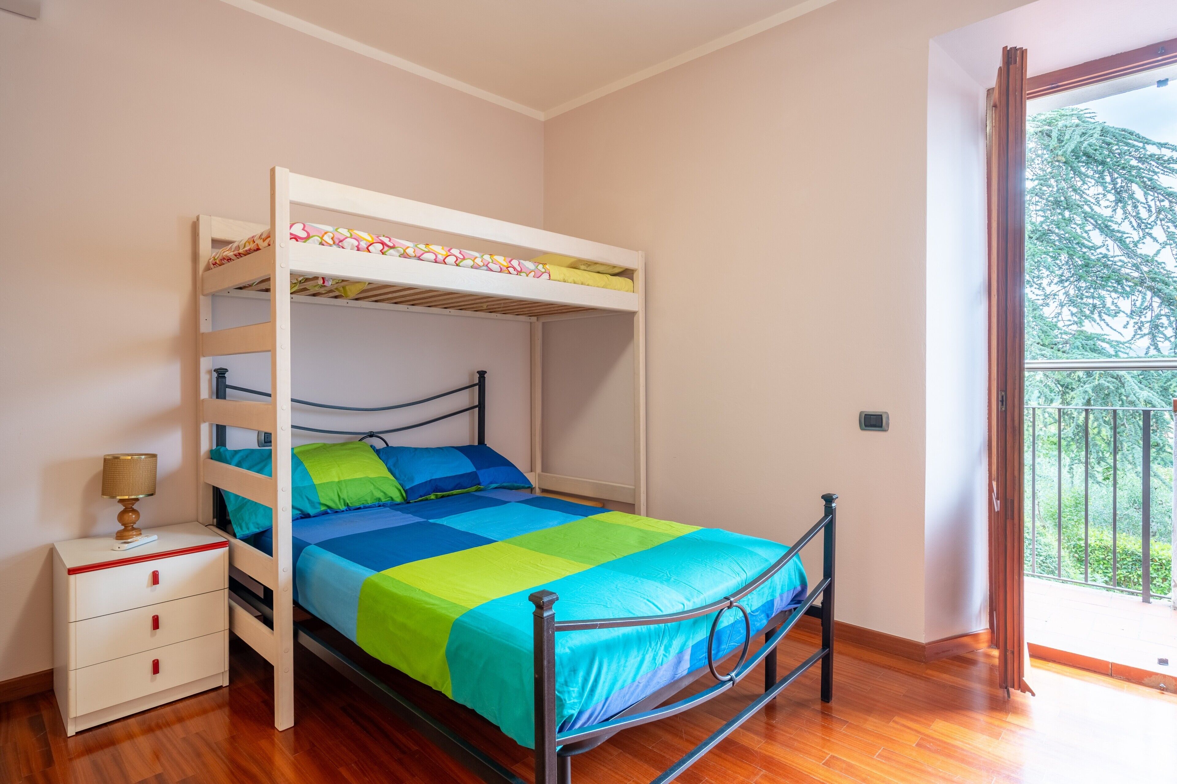 A bunk bed with a lower bed and a top bunk, a bedside table with drawers, and a window with a view of greenery.