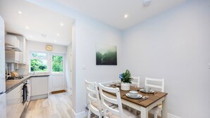 Et moderne køkken med hvide skabe, et spisebord dækket til fire personer, og udsigt til grøn natur udenfor.