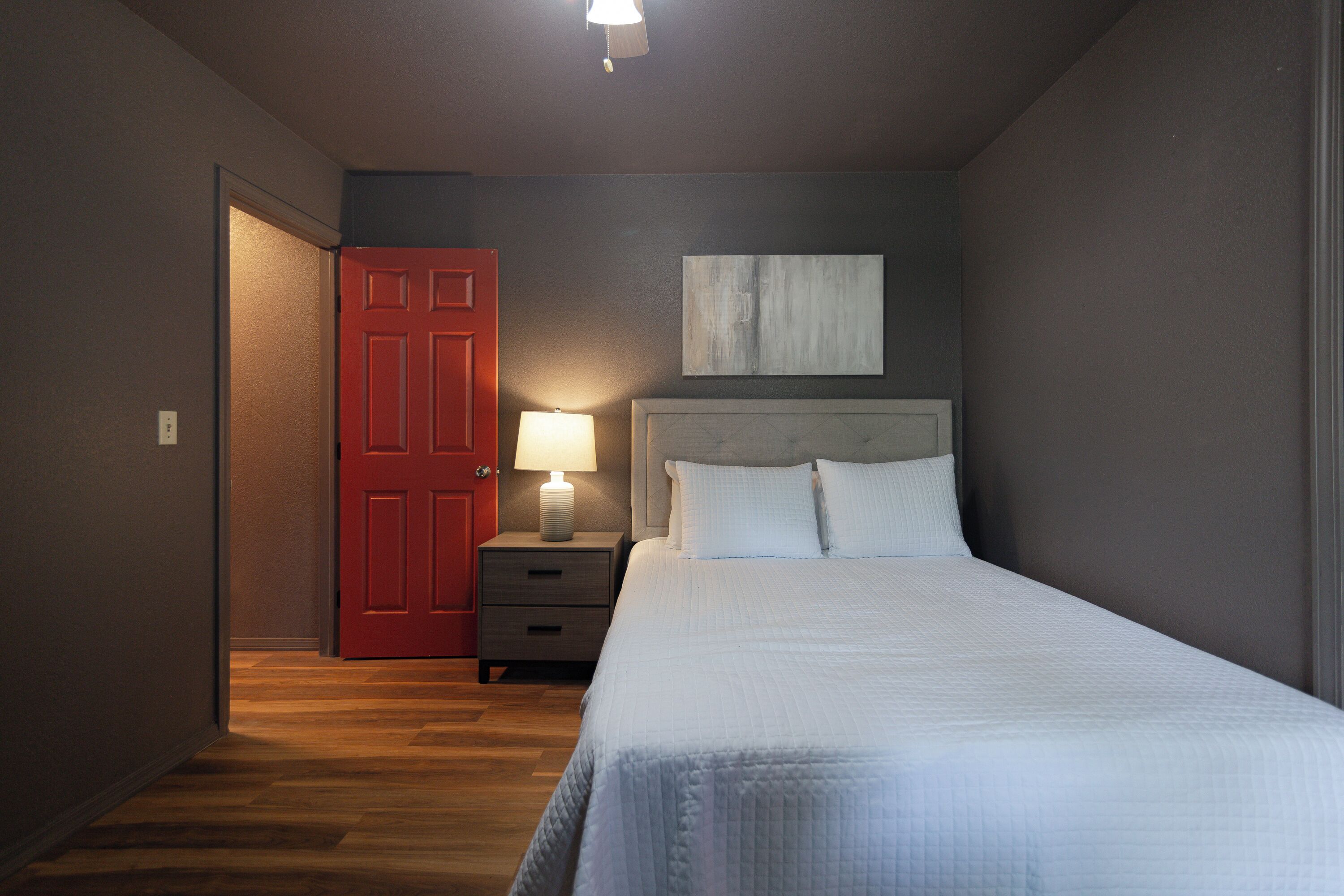 A bedroom with a bed, a nightstand with a lamp, a red door, and a painting on the wall.
