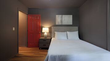 A bedroom with a bed, a nightstand with a lamp, a red door, and a painting on the wall.