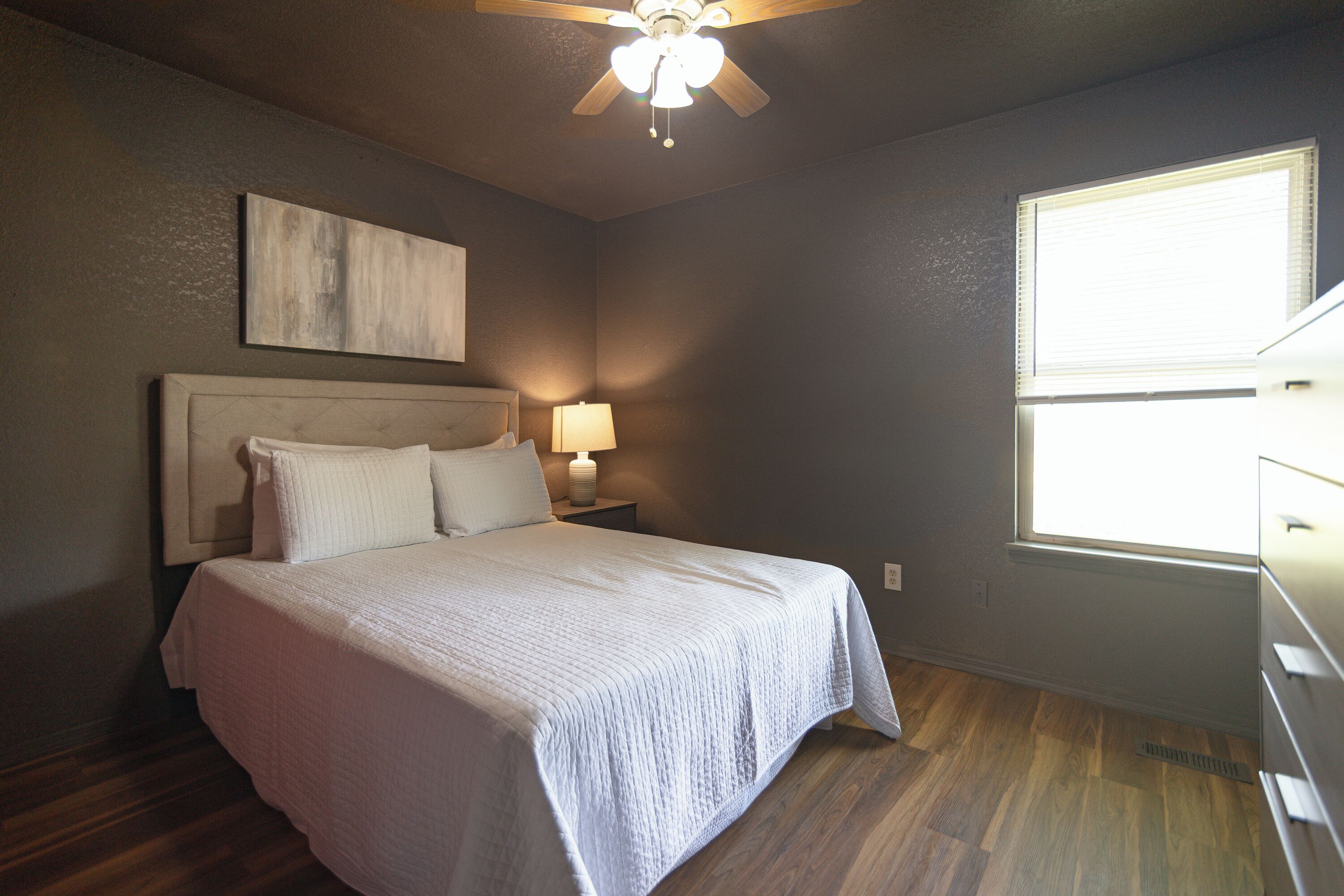 A bedroom with a bed, a nightstand with a lamp, a ceiling fan, and a window.