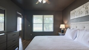 A bedroom with a bed, dresser, nightstand, lamp, and a window with blinds.