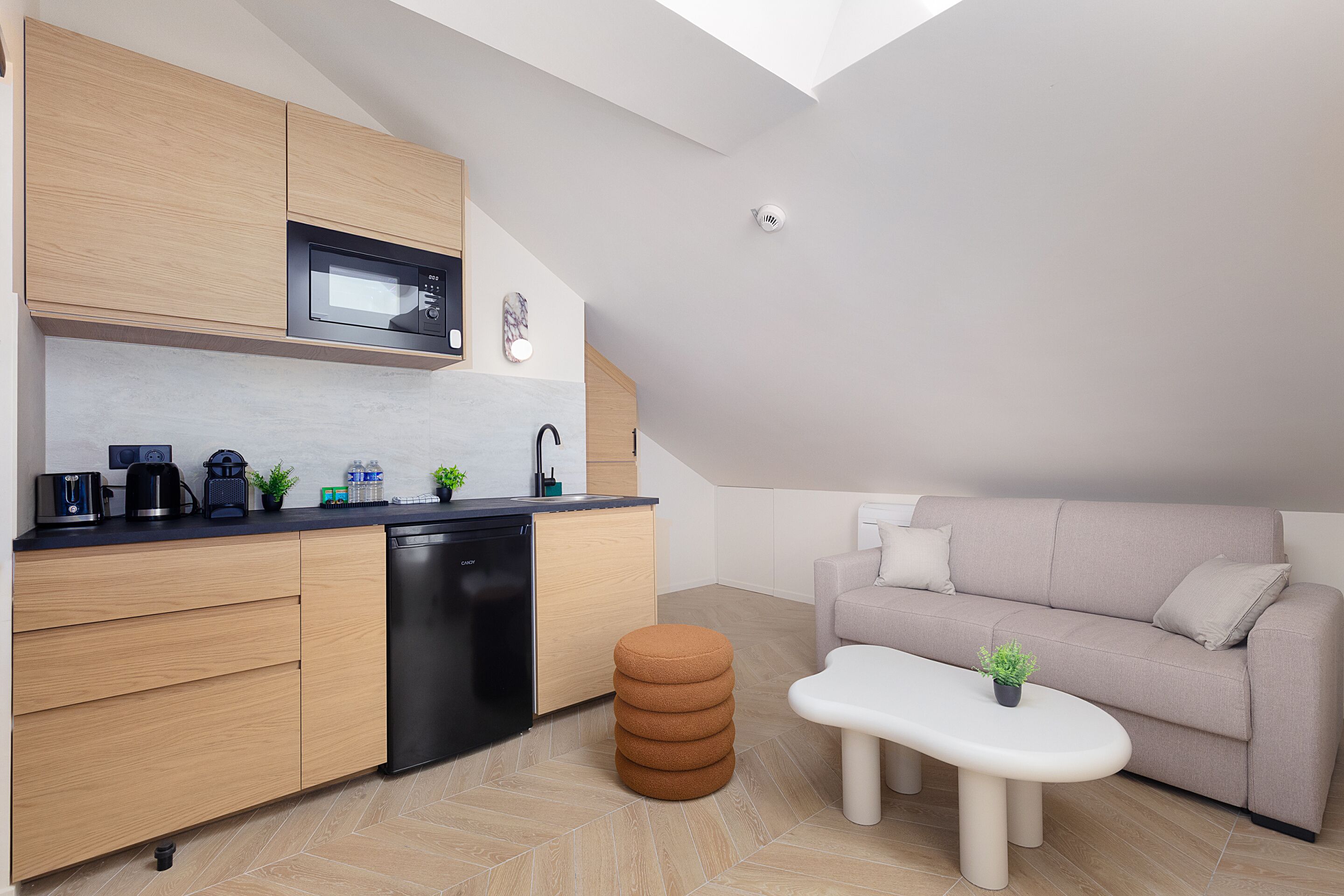 Une cuisine compacte dotée d’appareils électroménagers modernes, un coin repas avec un canapé et un pouf, et une lucarne bien visible.