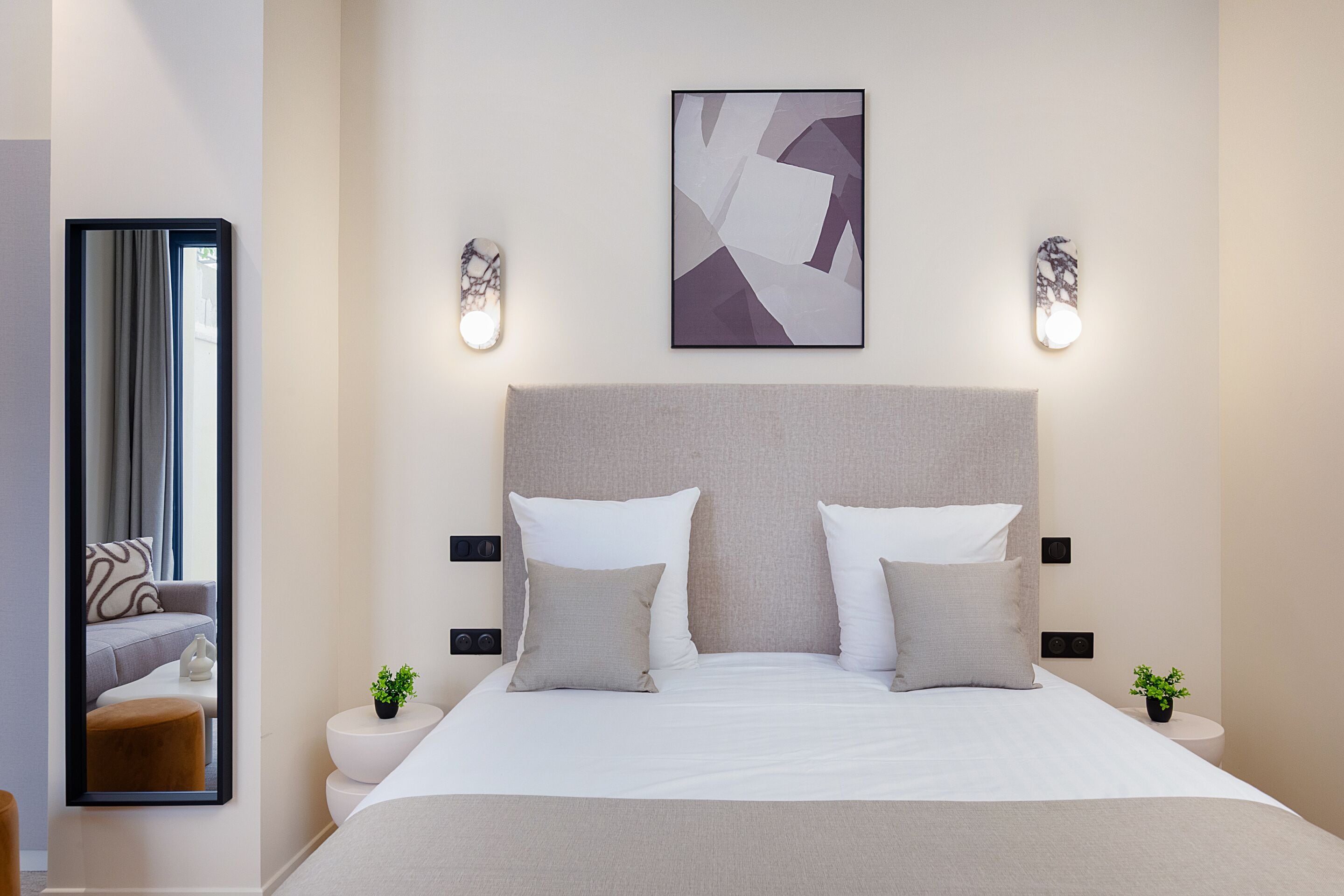 Una habitación de hotel moderna con una cama grande, dos mesitas de noche, un cabecero, una obra de arte abstracta colgada en la pared y un espejo que refleja parte de la habitación.