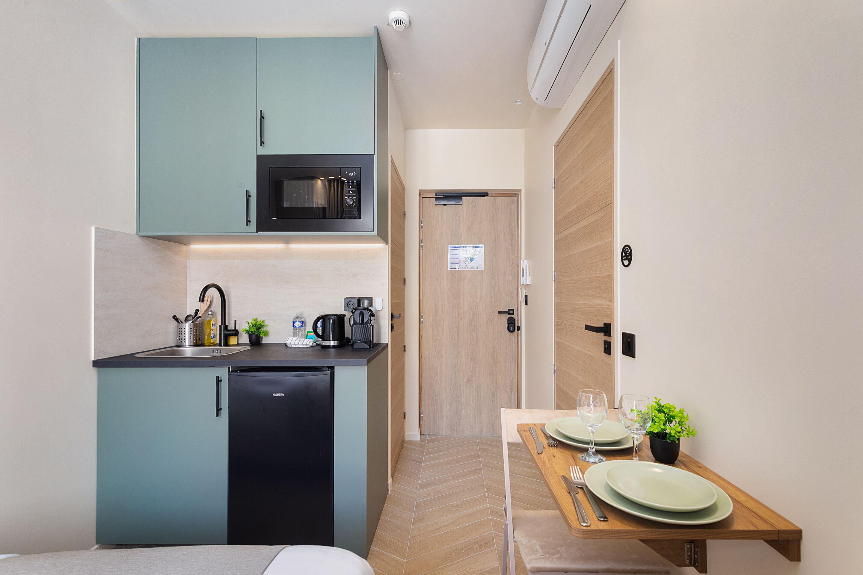 A compact kitchen with a microwave, sink, and coffee maker, a dining area with a table set for two, and a wooden door leading to another room.