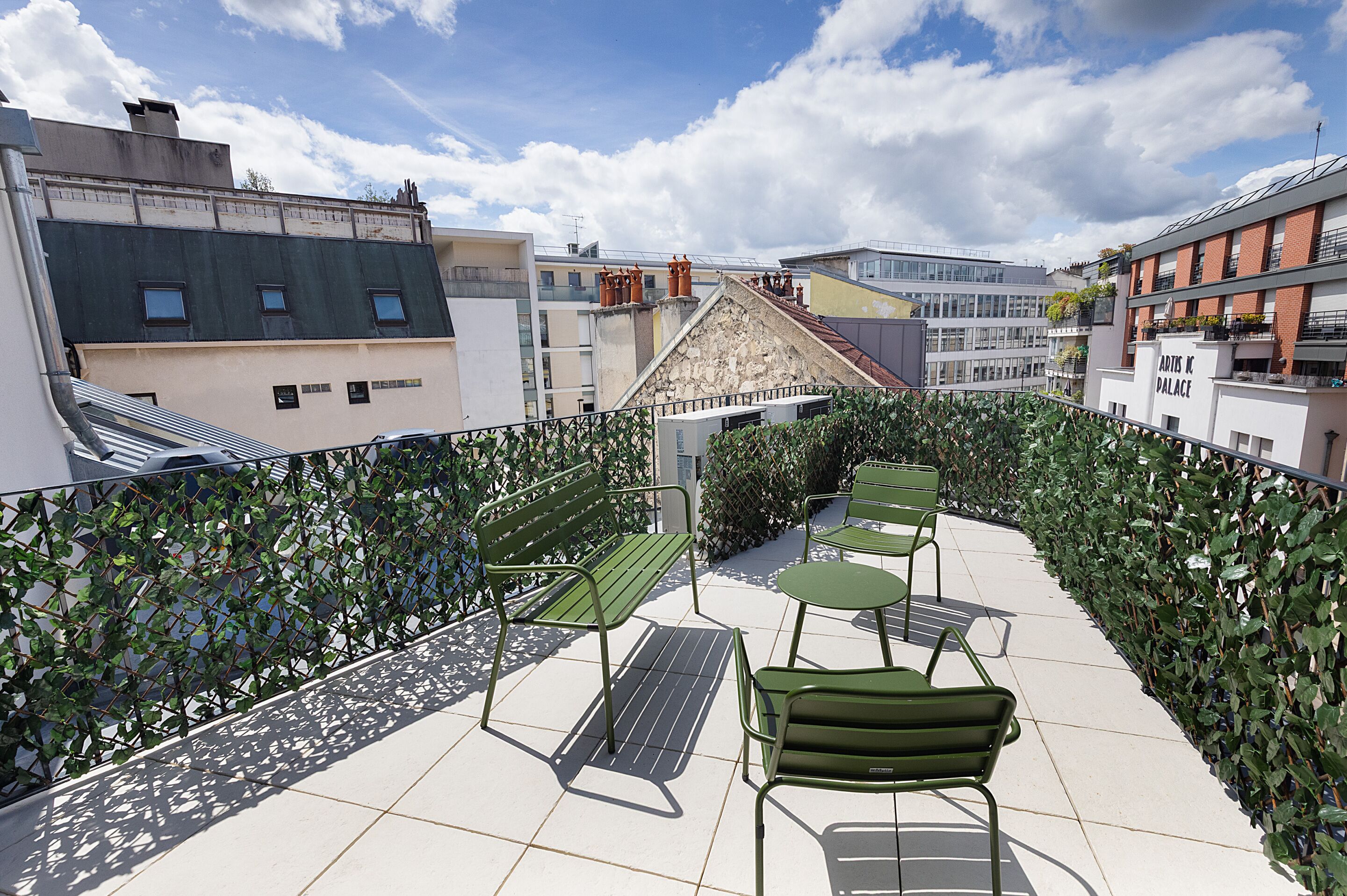 Una terraza en la azotea con mobiliario metálico verde, una barandilla de metal y vistas de edificios circundantes.