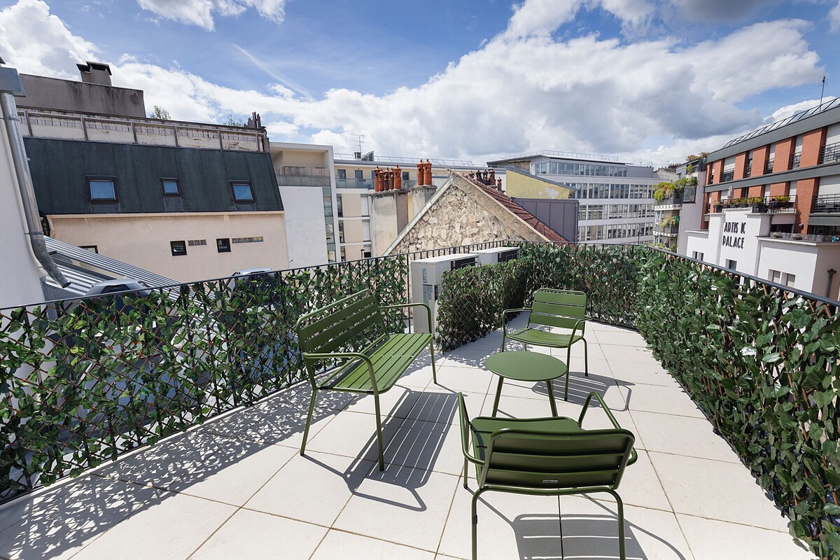 Una terraza en la azotea con mobiliario metálico verde, una escalera de metal y vistas de edificios circundantes.