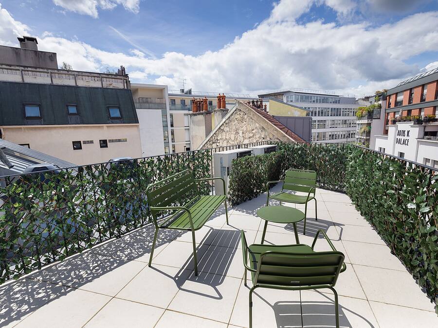 A rooftop terrace with green metal furniture, a metal staircase, and a view of surrounding buildings.