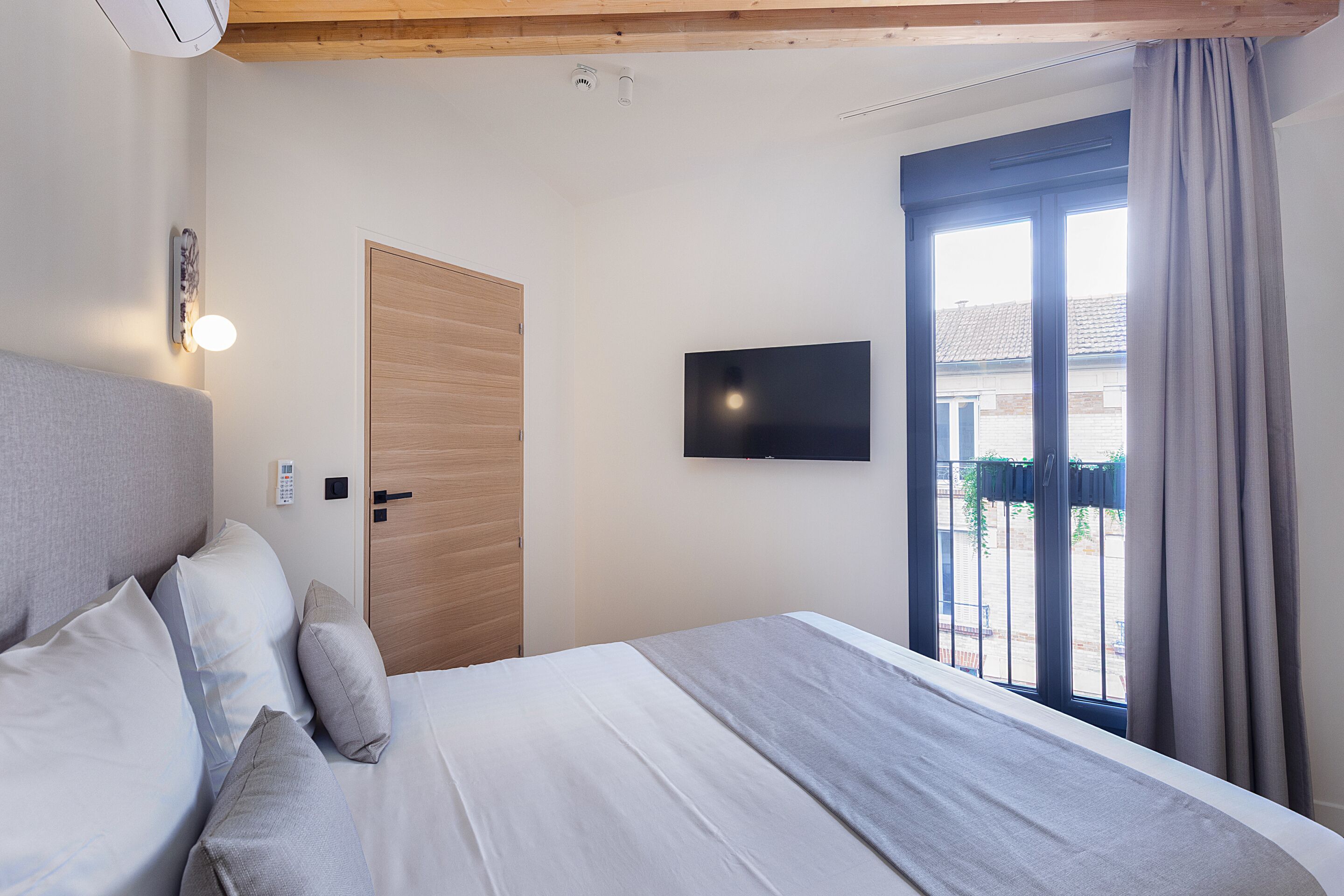 Een slaapkamer met een bed, een aan de muur bevestigde televisie, een deur en een raam met gordijnen.