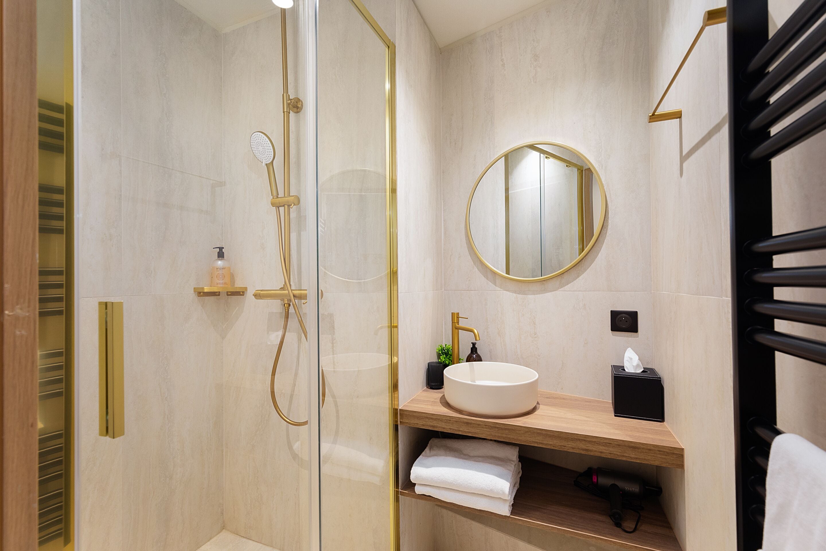 A modern bathroom with a glass-enclosed shower, a round mirror, a sink with a wooden countertop, and a towel rack.