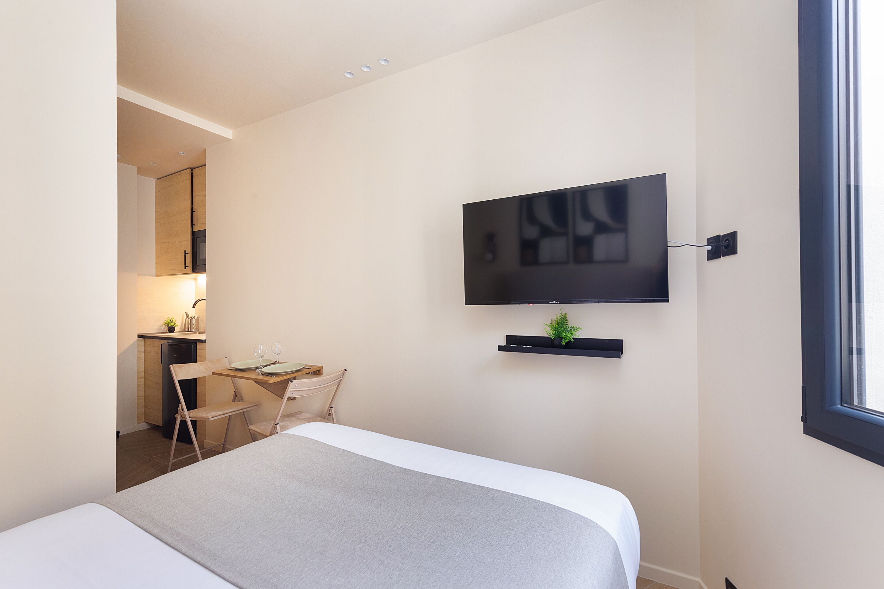 A modern hotel room with a bed, a desk with a chair, a television mounted on the wall, and a window.