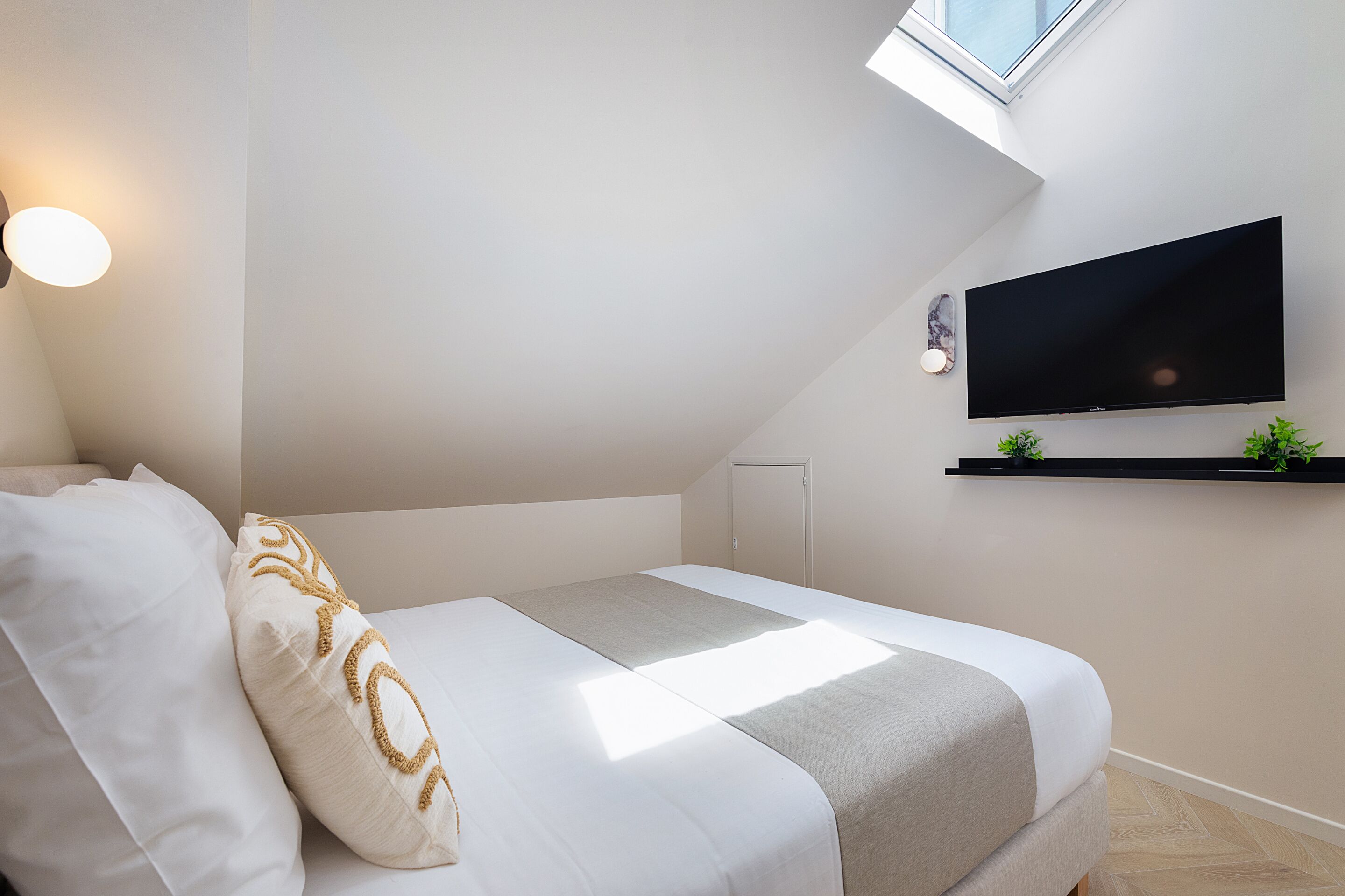 Un dormitorio moderno con una cama, un televisor instalado en la pared, un tragaluz y un estante pequeño con plantas.