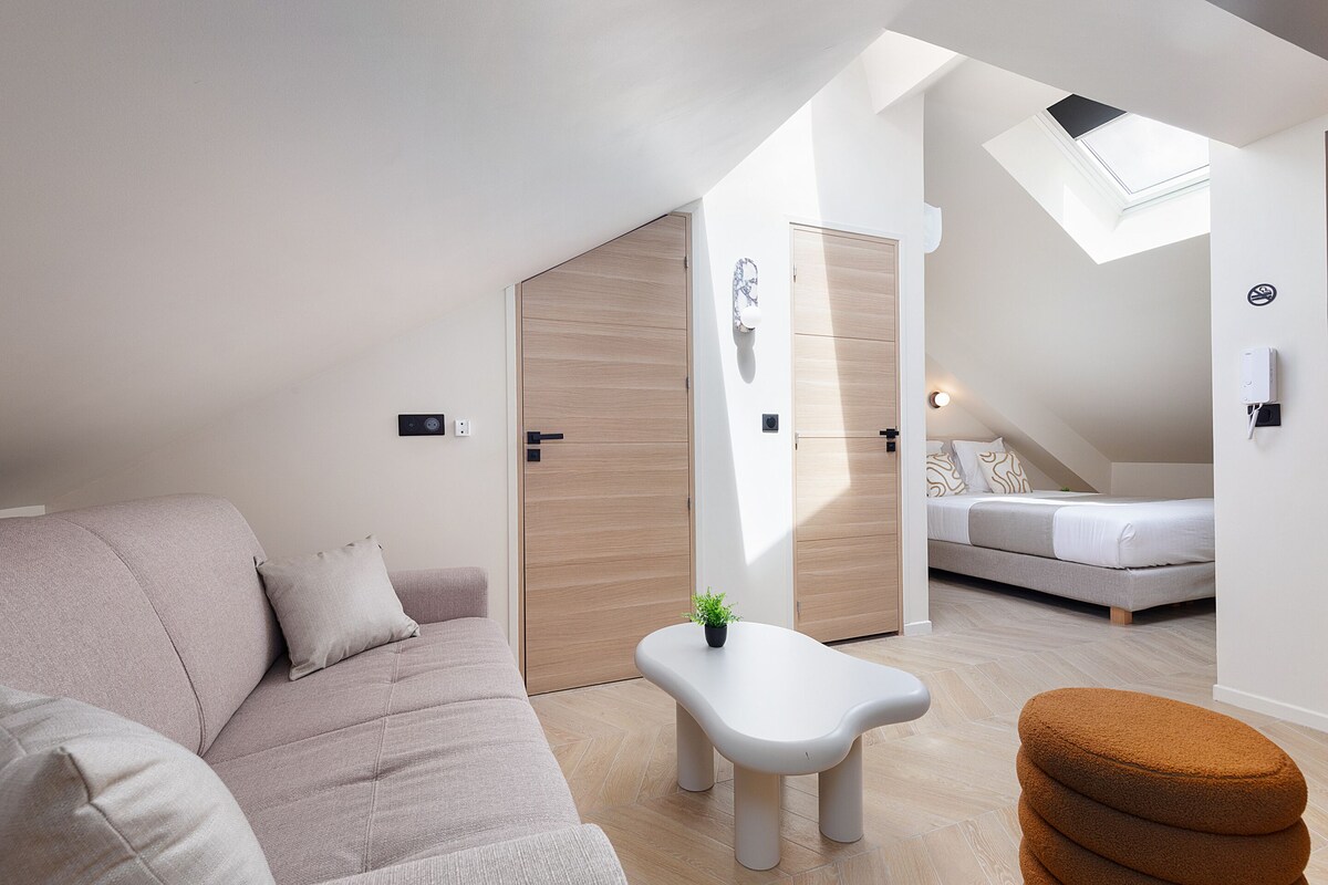 Un interior moderno con un sofá, una mesita con una planta, un pouf marrón y una cama al fondo.