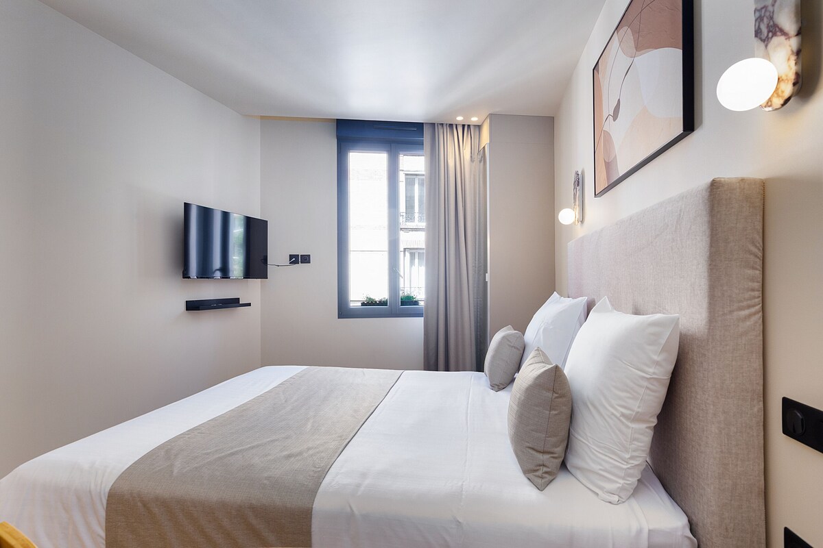 Una habitación de hotel moderna con una cama grande, un televisor de pantalla plana en la pared, una ventana con cortinas y una obra de arte enmarcada sobre la cama.