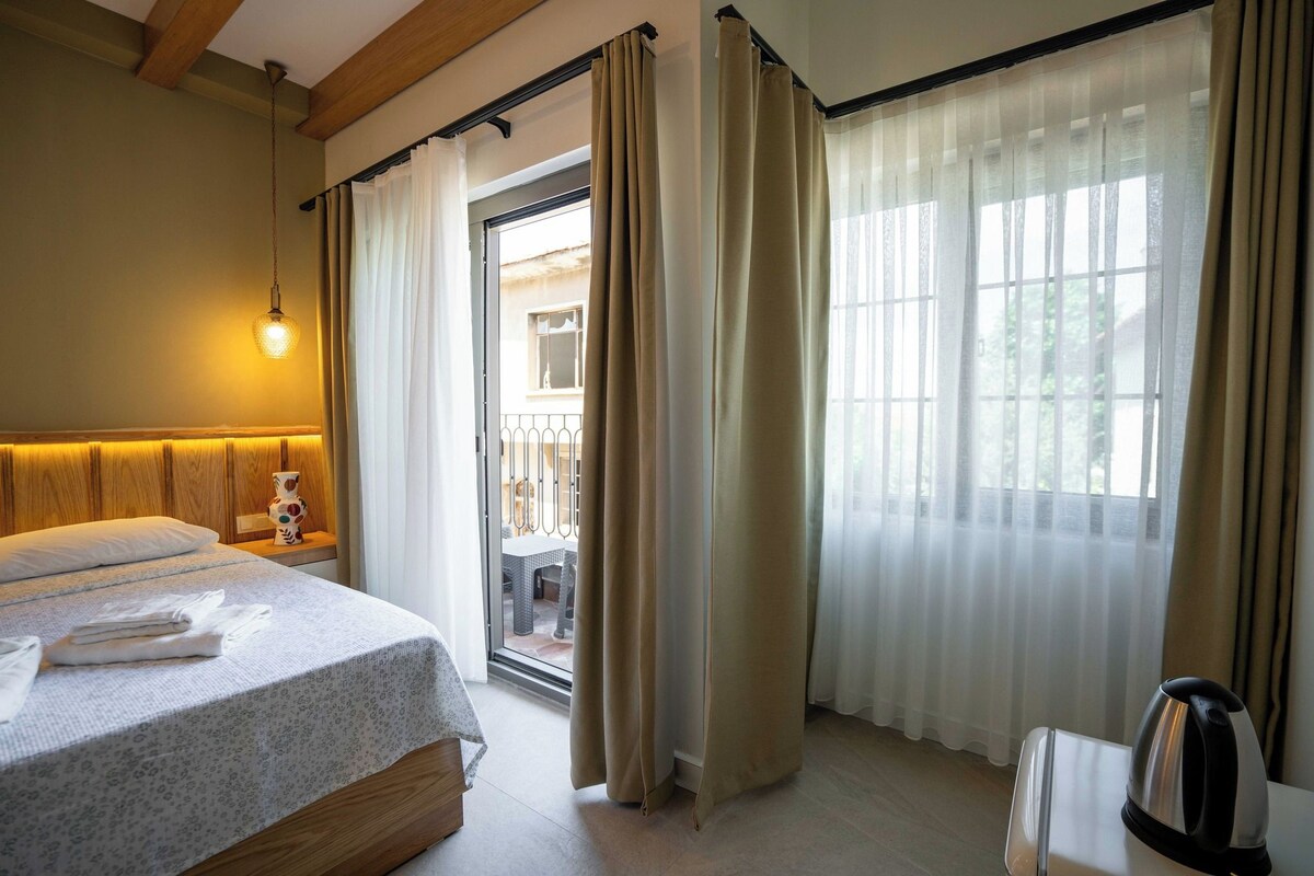 Habitación de hotel con cama, mesita de noche con lámpara, vista a través de una puerta de cristal y una tetera en el suelo.