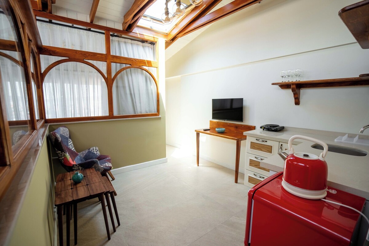 A compact living space with a television, a small table, a red kettle, and a window with curtains.