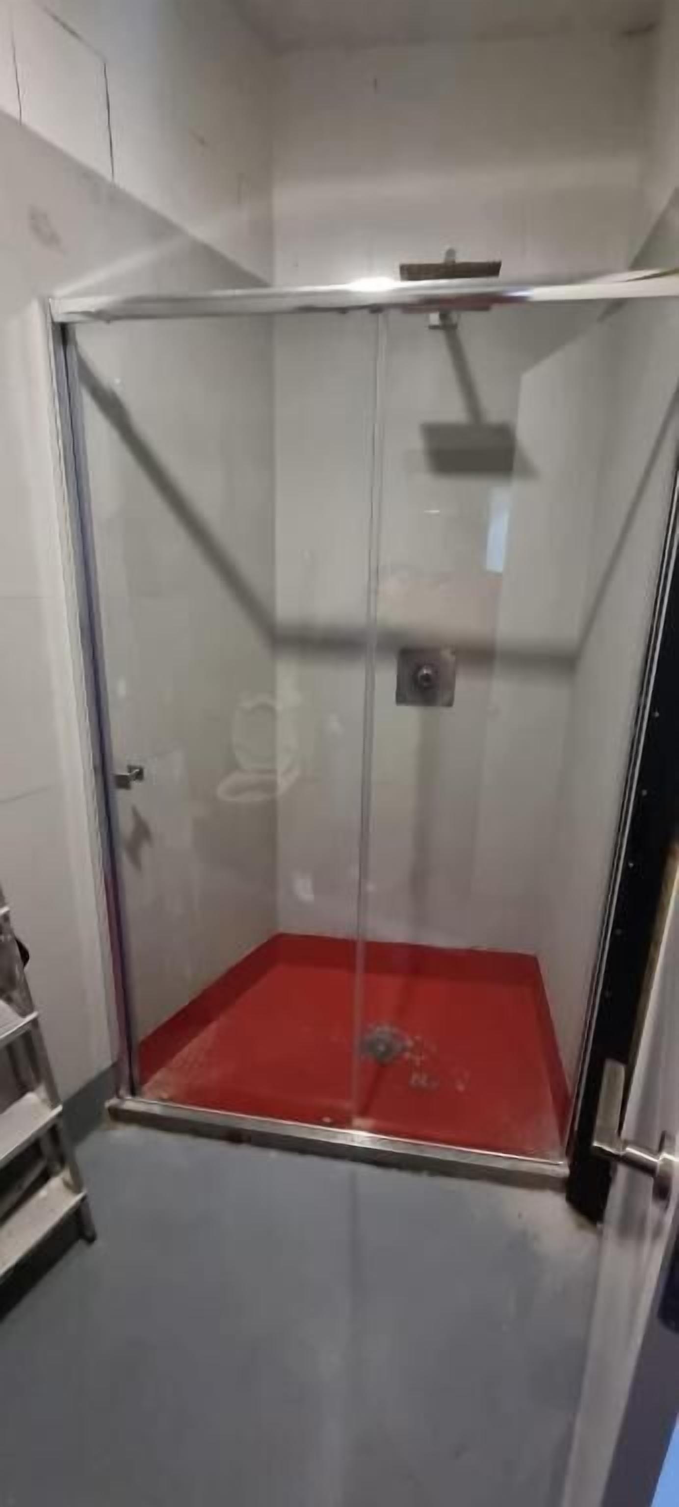 A glass-enclosed shower stall with a red floor, a visible showerhead, and a metal shelf.
