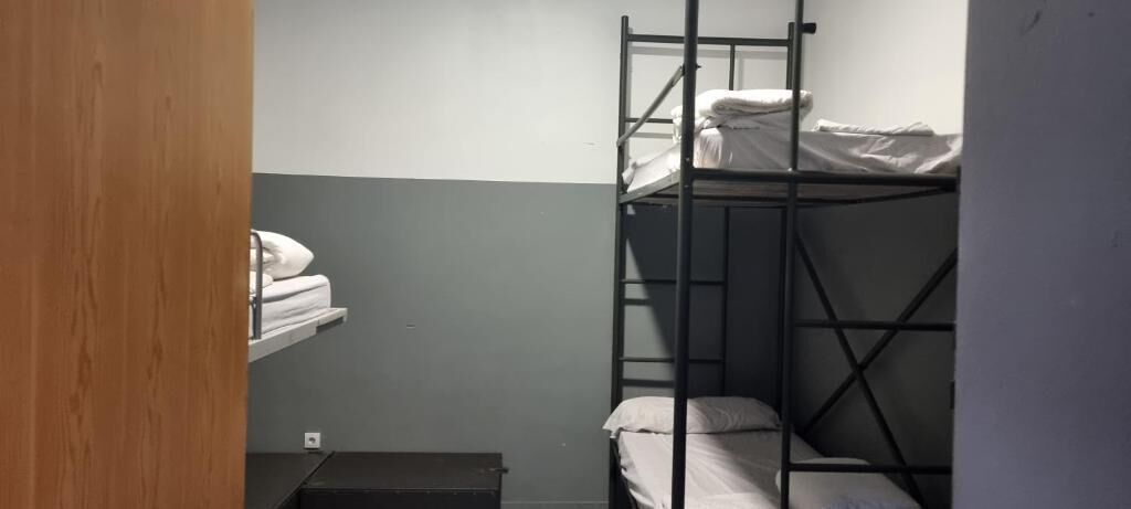 A bunk bed room with a shelf above it, a small bench, and a wall socket.