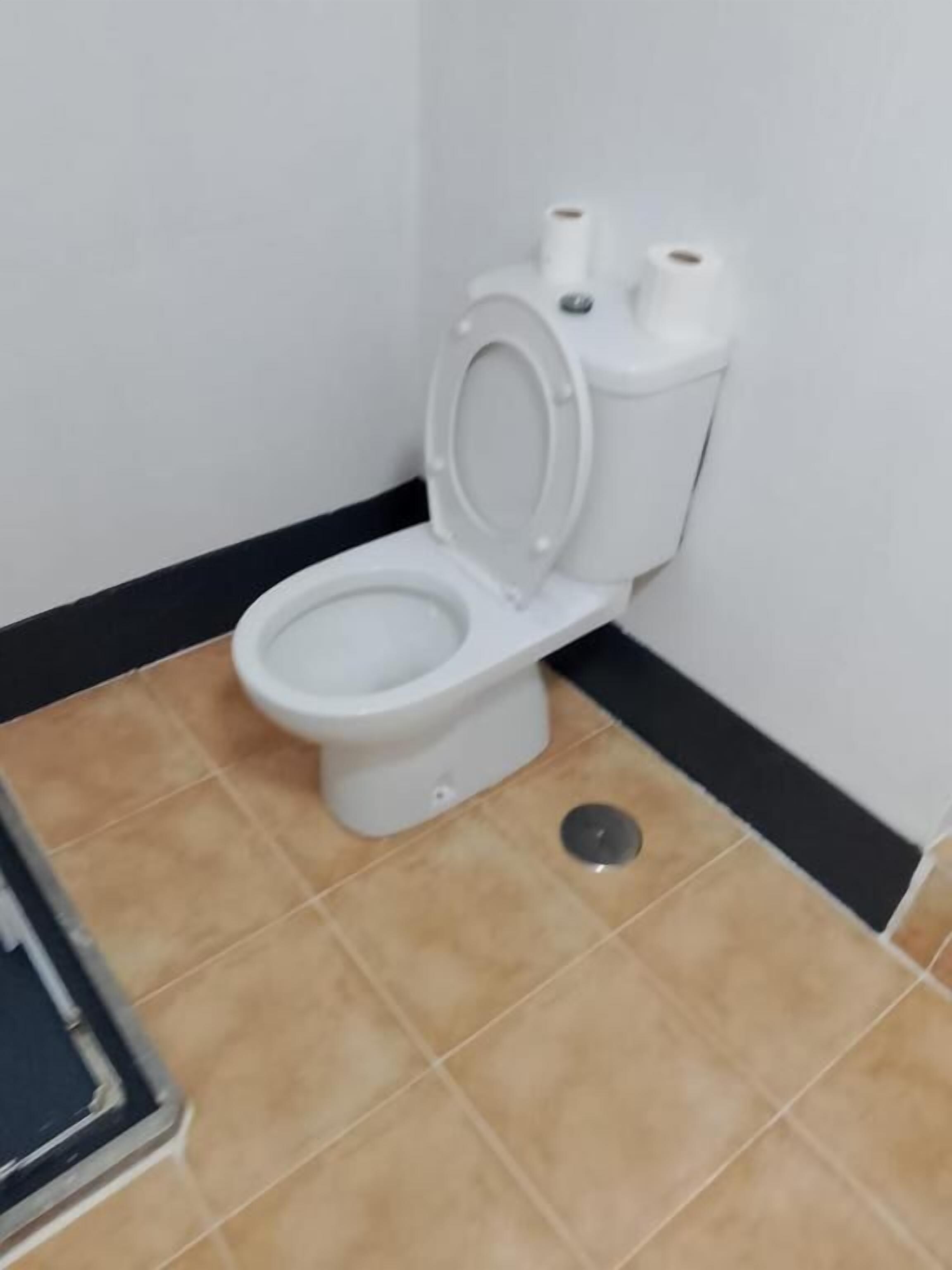 Inodoro estándar con tanque y asiento blancos, ubicado en una esquina de una habitación con piso de baldosas color beige y zócalo negro.