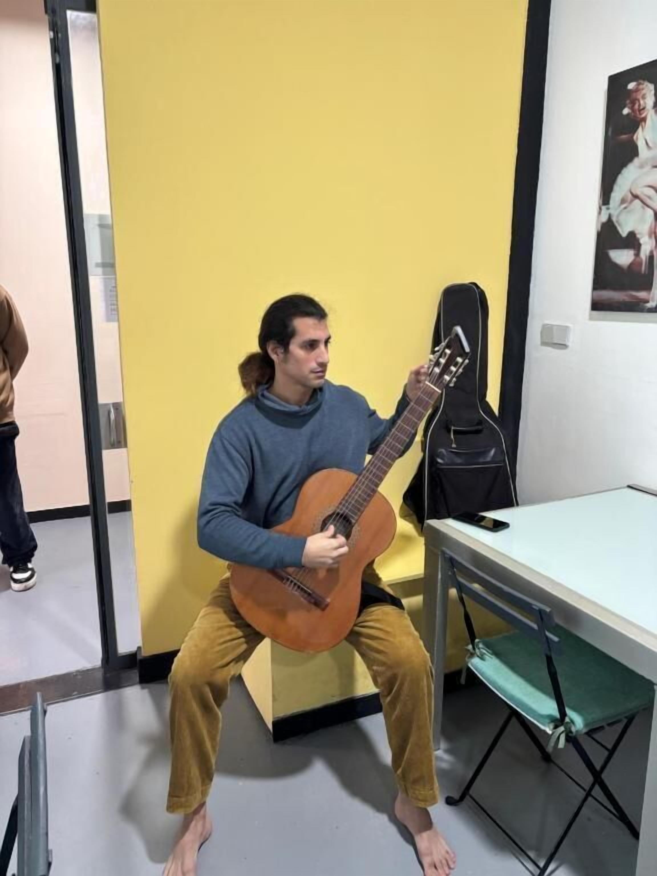 A person sitting on a stool playing a guitar in a room with a yellow wall and a white table.