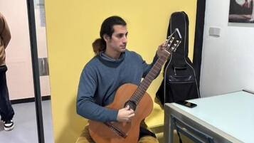 Una persona sentada en un taburete tocando una guitarra en una habitación con pared amarilla y mesa blanca.