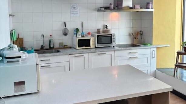 Une cuisine avec des meubles blancs, un grand îlot central doté de tabourets, un four à micro-ondes et un téléviseur qui diffuse une image d’une pièce.