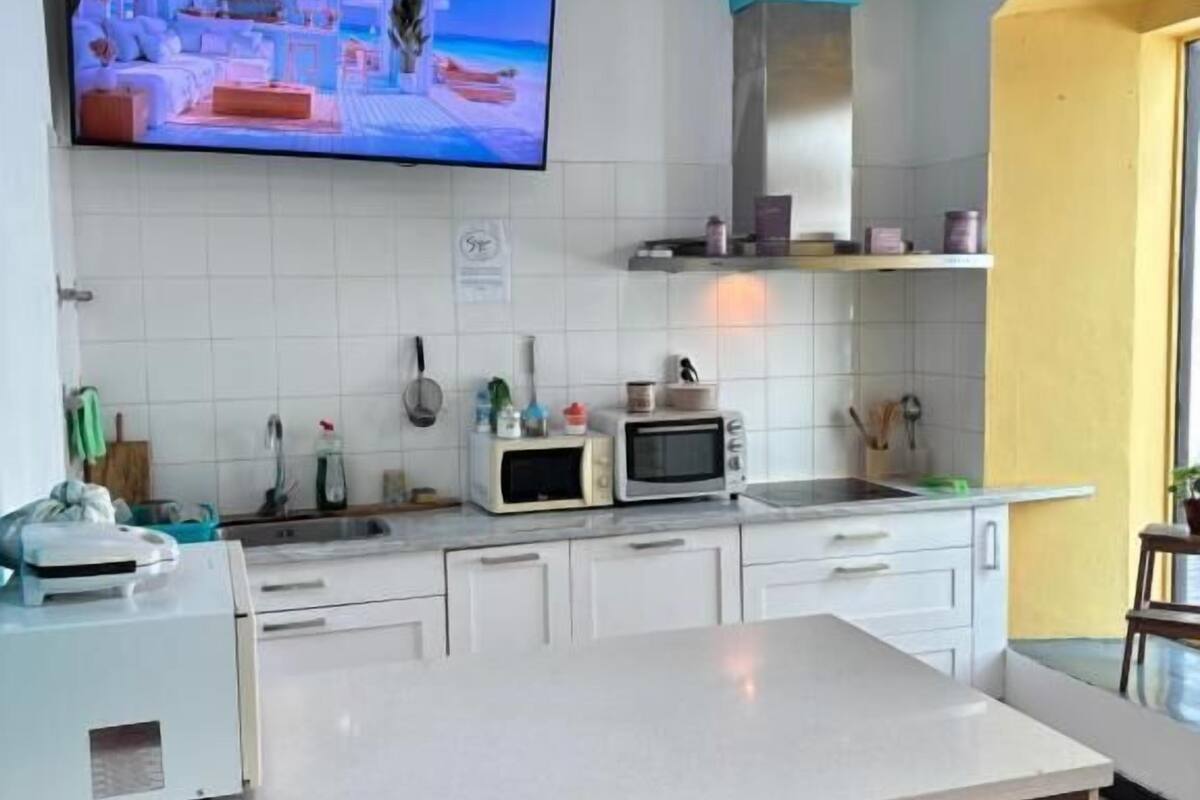 Una cocina con armarios blancos, una isla grande con taburetes de bar, un microondas y un televisor que muestra una habitación.