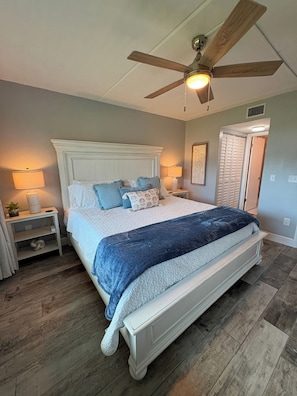 A bedroom with a bed, a ceiling fan, a nightstand, a lamp, and a door. - Beautiful 2br 2ba Ocean View Flat! Amazing Views and Location (St Augustine)