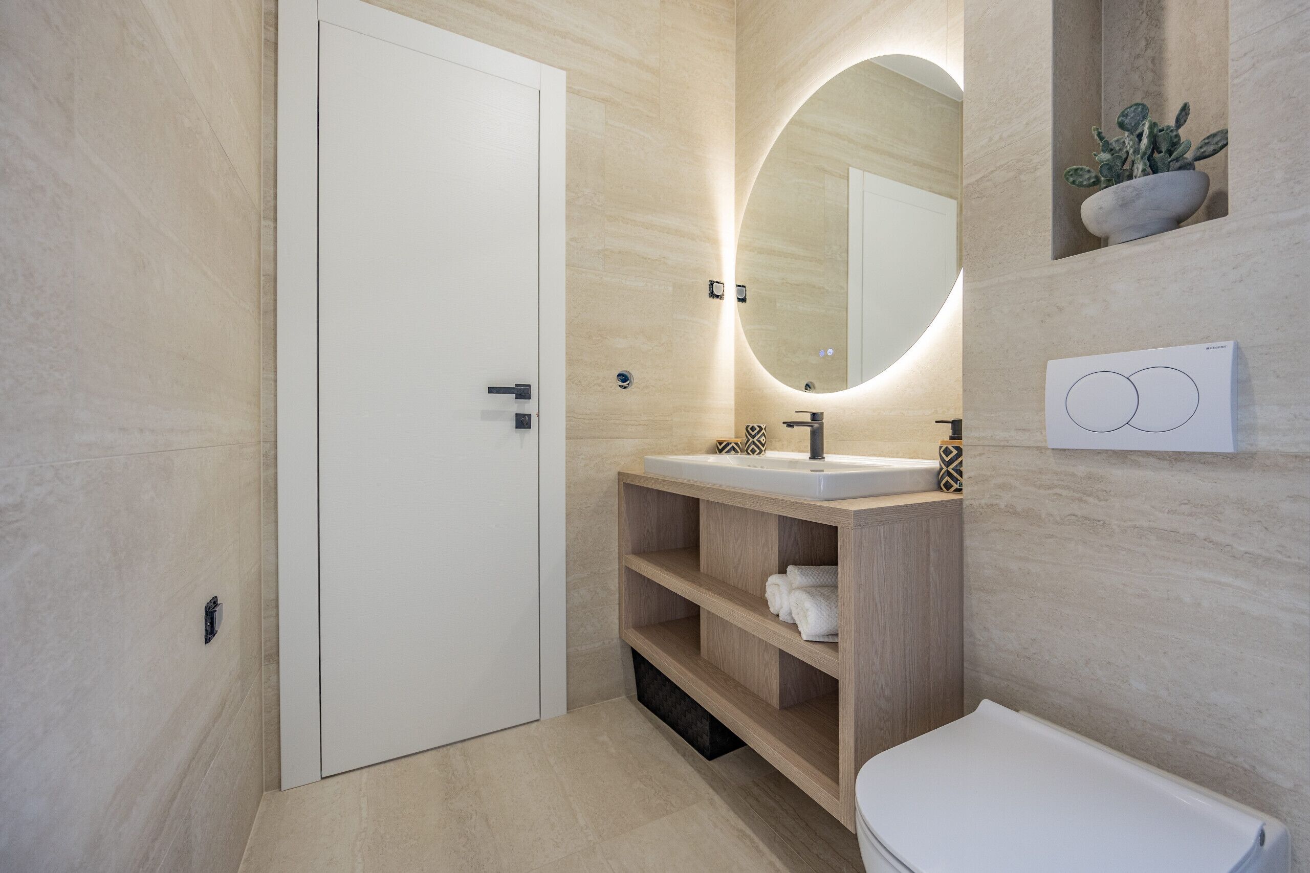 Een moderne badkamer met een witte deur, een ronde spiegel met verlichting, een wastafel met kraan en een plank met handdoeken.