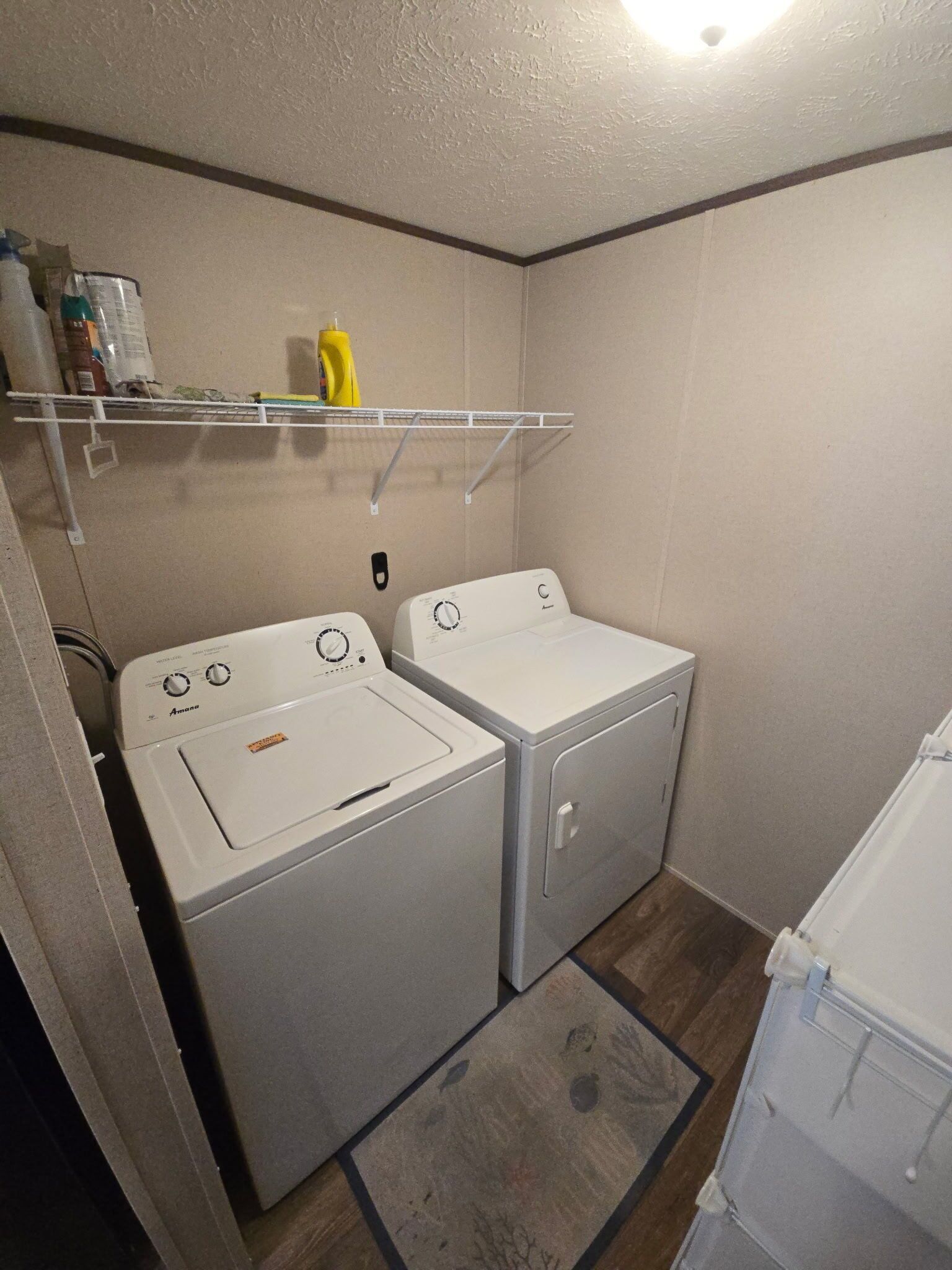 A laundry room with two white washing machines, a shelf with cleaning supplies, and a wooden floor.