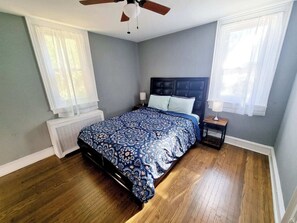 Ein Schlafzimmer mit Bett, Deckenventilator, Nachttisch und Fenster mit durchsichtigen Vorhängen.