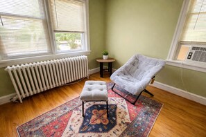 A corner room with a radiator, a grey armchair, a small table with a plant, and a patterned rug.