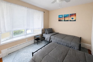 A bedroom with two beds, a window with blinds, a radiator, and a ceiling fan.