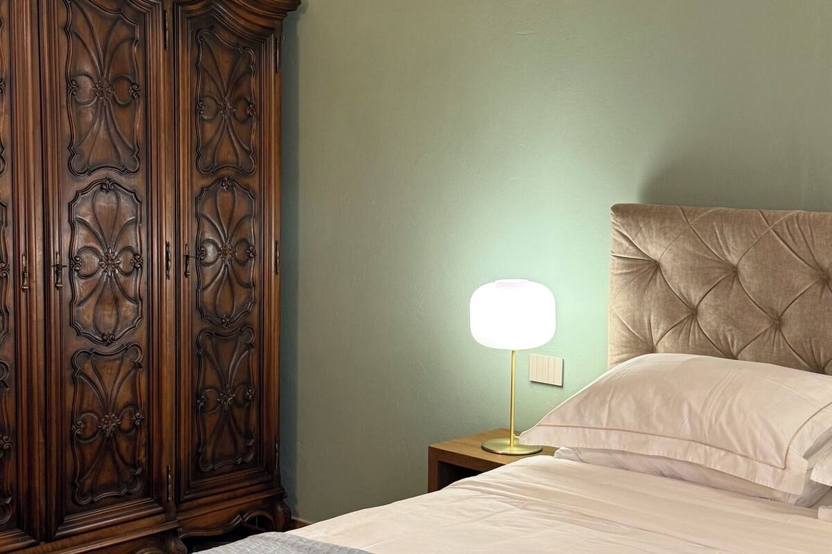A bedroom with a large wooden wardrobe, a bedside lamp, a bed with a tufted headboard, and a nightstand with a notepad.