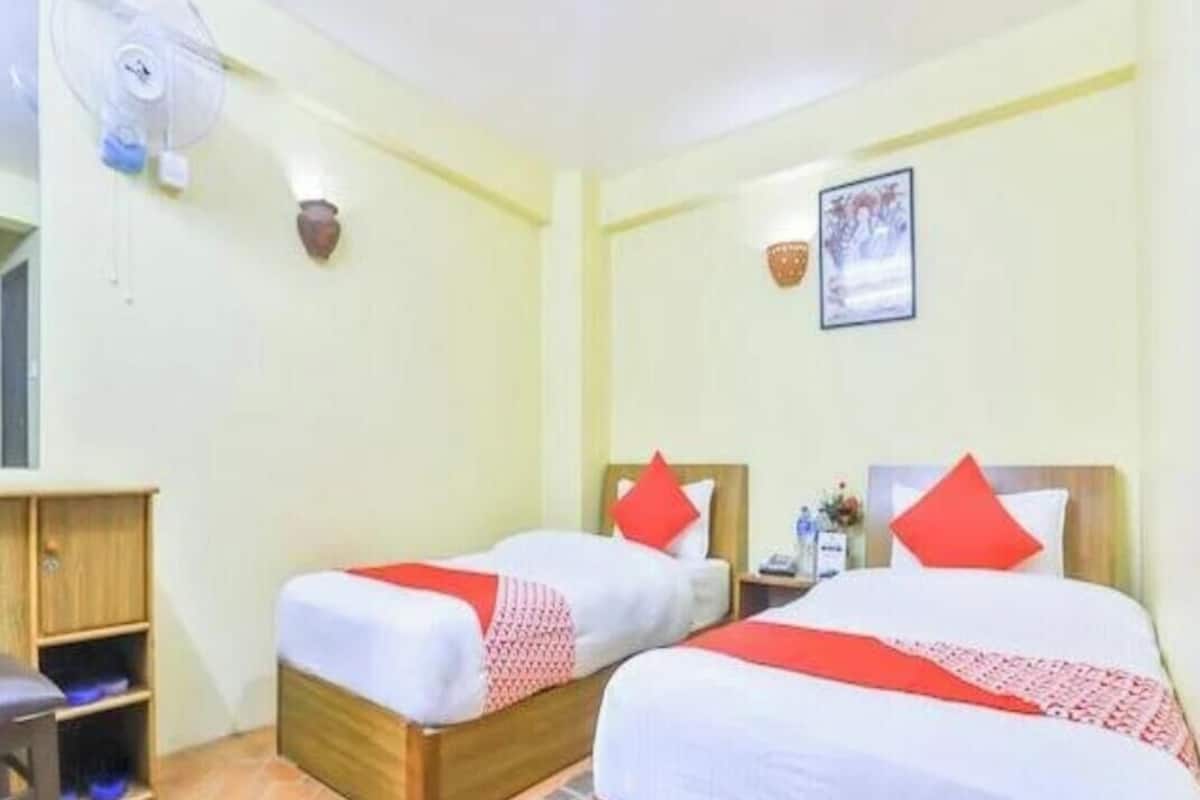 Habitación de hotel con dos camas, cabecera de madera, mesita de noche, ventilador de pared y un estante con objetos.